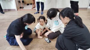 Toddler sits on a wooden floor as three adults kneel around, examining her foot during a small group activity; a phone with stickers lies nearby.