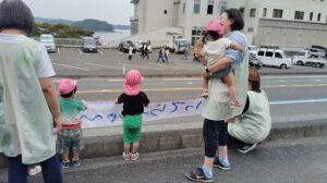 Children with pink hats and adults in pale vests hold a long banner along a road while bystanders and parked cars line the street, waterfront in distance.