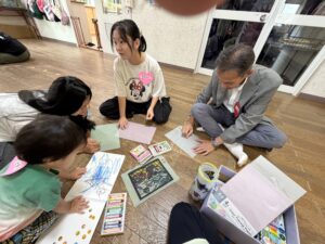 Group of children and an adult sitting on the floor, drawing with colored markers on paper in a classroom art activity area.