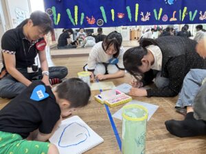 Group of children and adults sitting and kneeling on a classroom floor, drawing on papers with markers; colorful craft materials and a blue ocean-themed banner above them.