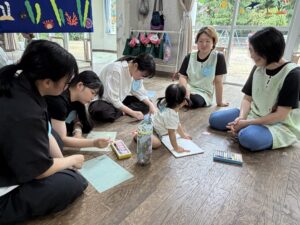 Group of adults and children seated on a wooden floor, drawing with markers and papers in a bright classroom filled with poster art.