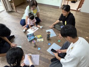 Group of children and adults sitting on a wooden floor, drawing with colored pencils and markers around papers and art supplies.