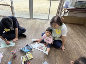 Adult woman and a man sitting on a wooden floor helping a young child draw with crayons and papers spread out, pencil cases nearby and a fish tank on a stand in the background.