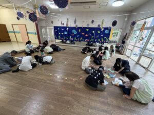 Group of adults and children sitting on the floor creating art, with a blue sea-themed backdrop and hanging decorations above.