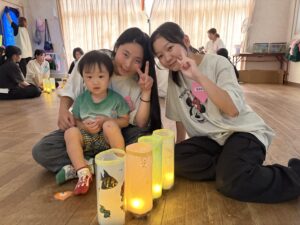 Three children pose for a photo indoors next to colorful paper lanterns with candles and other people in the background.