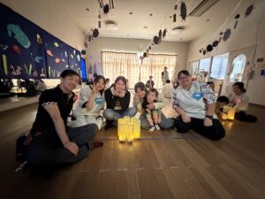 Group of adults and children sitting on the floor during an indoor activity, with ocean-themed wall art and lanterns lit in the foreground.