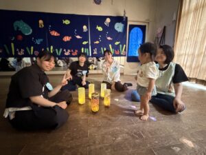 Group of kids sitting on a wooden floor in front of a blue ocean mural with sea creatures and coral, holding up peace signs during a classroom activity.