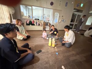 People sit in a circle on a wooden floor, a baby with an adult, and yellow cups on the ground for a group activity in a classroom-like room.