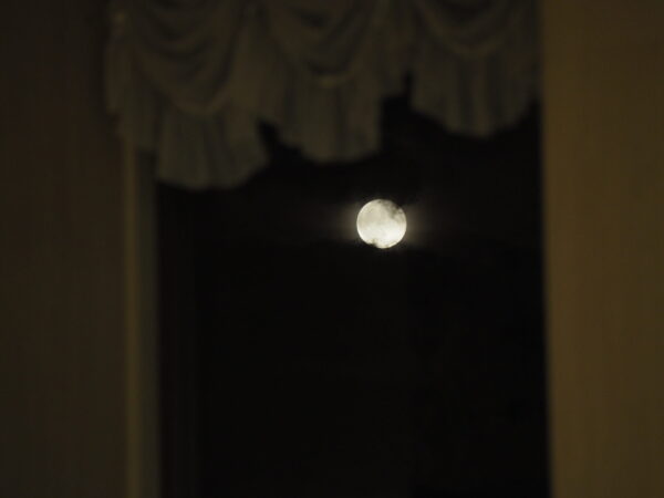 Moon visible in a dark night sky through a window with indistinct curtains or drapes.