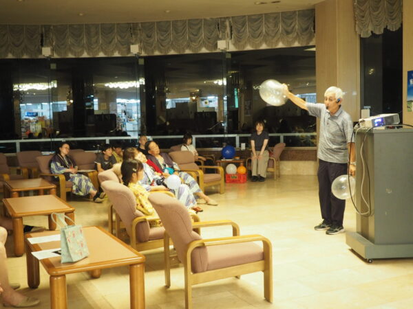 Older man with a headset and microphone speaking to a seated audience in a lounge, holding a large clear balloon; projector on a podium to the right.