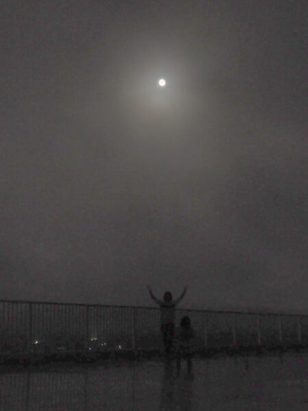 Moon with a halo glowing in a cloudy night sky above a distant city skyline at dusk.