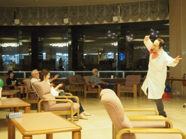 Entertainer in a white coat and red bow tie performing a lively gesture for seated audience in a lounge area with glass walls.