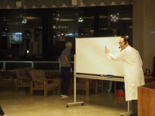 Instructor in a white coat with a headset writing on a whiteboard during a class in a dimly lit lecture room.