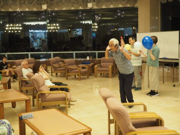 Elderly man with a headset gestures as others hold colorful balloons near a whiteboard in a lounge.